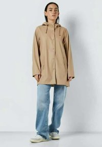 Lys beige regnfrakk med hette, frontlukk med trykknapper og to sidelommer. Kombinert med lyseblå vid bukser og casual sneakers.