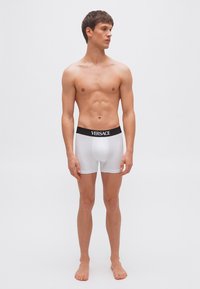 Νεαρός άνδρας που στέκεται ξυπόλυτος φορώντας λευκά εσώρουχα boxer της Versace με μαύρη μέση, κοιτάζει προς τα δεξιά του πάνω σε απλό φόντο.