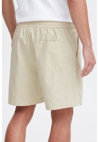 Beige shorts with elastic waistband, single back pocket, and textured fabric. Features a straight cut and stitched hem.