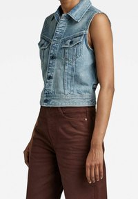 Ljusblå denim ärmlös jacka med framfickor, knäppning och cropped design. Tyget har en urtvättad textur.