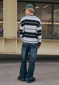 Camicia a maniche lunghe a righe bianca, nera e rosa chiaro, abbinata a jeans in denim blu a gamba larga e scarpe nere. Cappellino verde in testa.