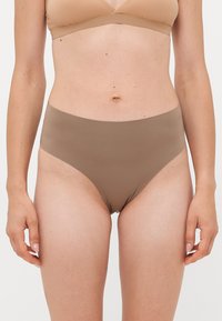 Woman wearing high-waist seamless brown underwear and a beige bra, standing with arms relaxed by her sides on a white background.