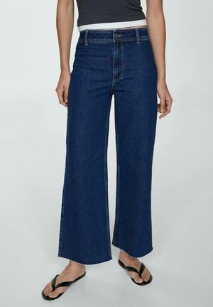 Personne portant un jean large bleu foncé court avec des ourlets effilochés, des tongs noires et un haut noir sans manches sur un fond uni.