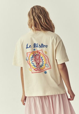Person with wavy hair wearing a cream t-shirt featuring a red lobster and text "Le Bistro" on the back, paired with a pink striped skirt.