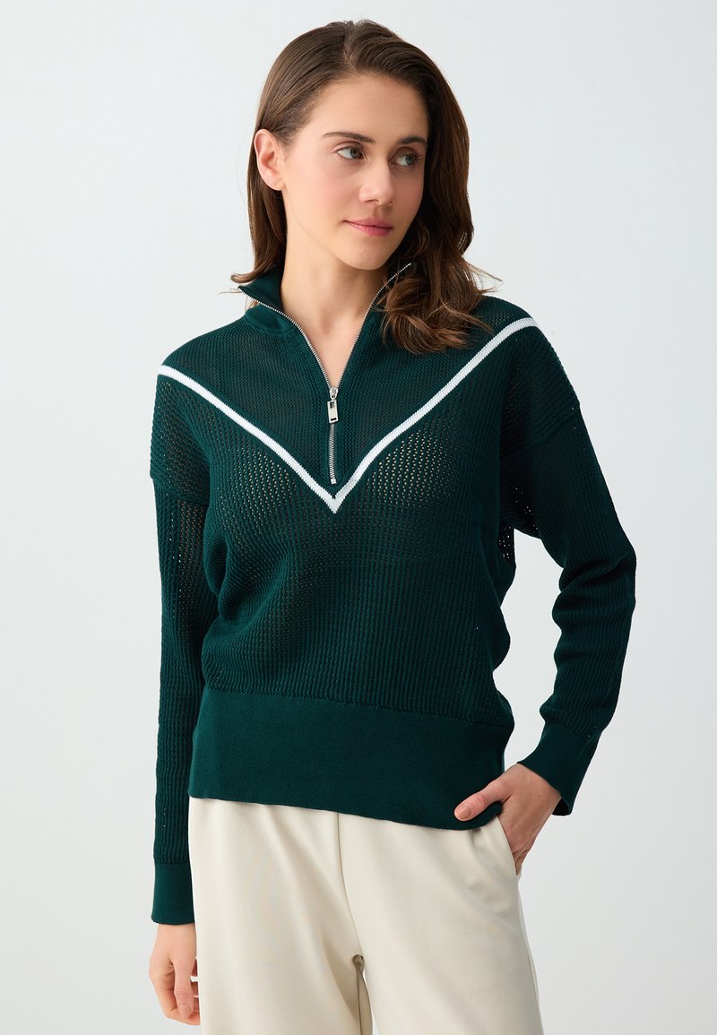 Femme aux cheveux bruns portant un pull en maille vert foncé avec des rayures chevrons blanches et une fermeture éclair à l'avant, associée à un pantalon beige clair.