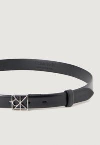 Czarny skórzany pasek z geometryczną klamrą w kolorze srebrnym, z wytłoczonym logo Calvin Klein na pasku. Gładka tekstura, prosty design.