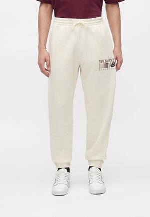 Personne portant un pantalon de survêtement New Balance crème avec logo sur la cuisse gauche, des baskets blanches et une chemise bordeaux visible de la taille vers le bas.