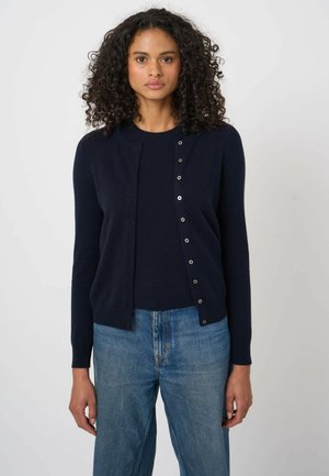 Kobieta z kręconymi włosami, ubrana w granatowy kardigan na guziki nałożony na pasujący sweter oraz niebieskie dżinsy, stojąca na jednolitym tle.