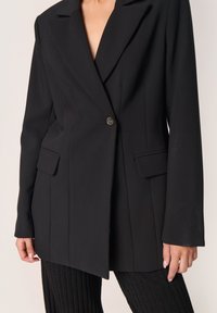 Schwarzer Blazer mit Reverskragen, Ein-Knopf-Verschluss, zwei Vordertaschen und strukturierten Schultern. Der Stoff hat eine glatte Textur.