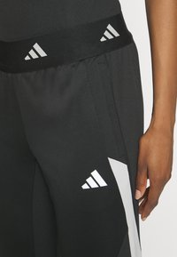 Zwarte sportbroek met een brede zwarte tailleband, voorzien van witte Adidas-logo accenten en lichtgrijze zijkanten. Gladde, nauwsluitende textuur.