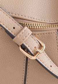 Bandoulière de sac en cuir beige texturé avec boucle dorée et fermeture éclair assortie dorée sur une surface en cuir avec des coutures visibles.