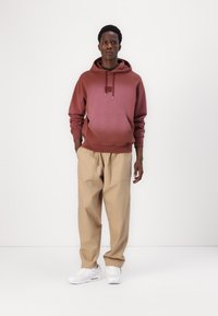 Maroon hoodie with a kangaroo pocket and adjustable drawstrings, paired with loose beige trousers and white sneakers on a plain background.