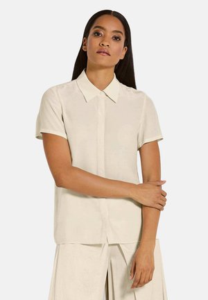Blouse beige à manches courtes avec un devant boutonné, col pointu et texture lisse, présentant une coupe droite et une coupe décontractée.