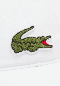 Boné de beisebol branco com um logótipo de crocodilo bordado em verde e vermelho, com detalhe texturizado, contornado em costura preta.