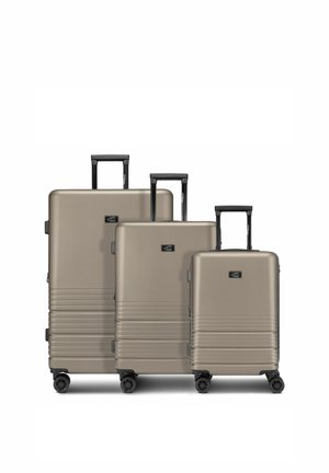 Tre valigie rigide beige di dimensioni diverse con manici neri estesi e quattro ruote girevoli, disposte fianco a fianco su sfondo bianco.