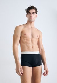 Zwarte boxershorts met een witte Calvin Klein-logo tailleband. De stof lijkt glad en elastisch. Het model heeft een gespierd lichaam en draagt geen shirt.