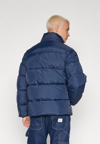 Mørkeblå puffjakke med højt krave, har vandret syninger, en blank finish og en løs pasform. Bæres med blå jeans.