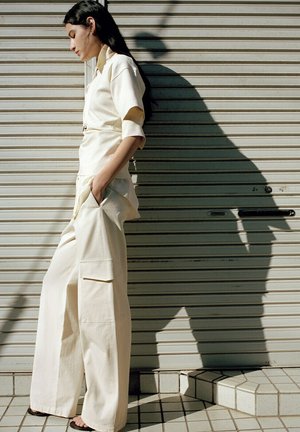 Donna in completo crema con pantaloni ampi e maniche arrotolate, in piedi di lato contro una serranda metallica, mani in tasca, che proietta un'ombra netta.