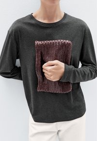 Camicia a maniche lunghe grigia indossata mentre si tiene una borsa a tracolla in pelle intrecciata di un rosso scuro. La borsa presenta un motivo a treccia con texture.