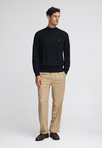 Svart lätt turtleneck-tröja med ribbat manschetter, i kombination med beige skräddarsydda byxor och bruna läderskor. Subtil logotypdetalj på bröstet.