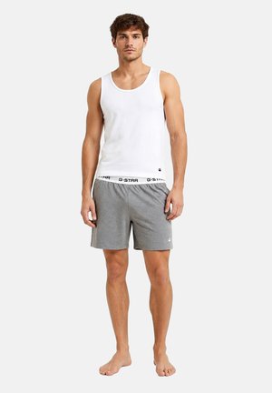 Man staat op blote voeten, draagt een wit mouwloos hemd en grijze korte broek met een elastische band met merknaam tegen een effen achtergrond.