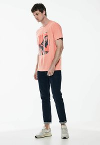 Triko s krátkým rukávem v korálové barvě s grafickým potiskem; spojené s tmavě modrými džíny a šedými teniskami; jemný materiál, neformální design.