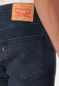 Sötétkék farmernadrág bőr címkével, amin a "Levi Strauss & Co." felirat található a hátsó részen. A nadrágon jellegzetes piros címke és ívelt zsebfektetések láthatók.