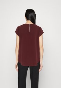 Burgundy kortermet topp med buet kant og glidlåsdetalj bak. Stoffet virker glatt og lett, kombinert med sorte bukser.