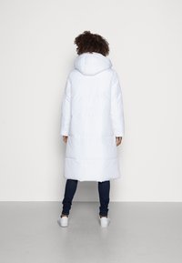 Abrigo acolchado largo, blanco, con capucha, que presenta secciones acolchadas horizontales y un dobladillo ligeramente redondeado. Combinado con jeans oscuros y zapatos blancos.