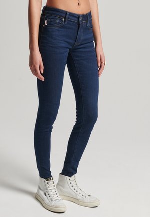 Jean slim bleu foncé porté par une personne debout, associé à des baskets montantes blanches sur un fond uni.