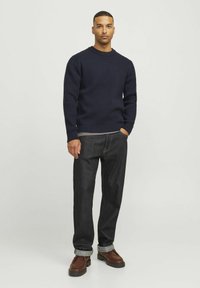 Dunkelblauer, gestrickter Pullover, lockere Passform, gerippte Textur. Kombiniert mit dunklen Jeans, am Knöchel umgeschlagen, und braunen Lederschuhen.