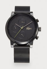 Montre chronographe noire avec un bracelet en maille texturée, trois sous-cadrans et des accents verts lumineux. Logo Lacoste visible sur le cadran.