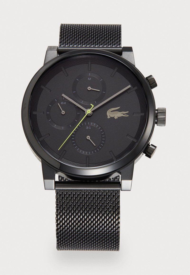 Montre chronographe noire avec un bracelet en maille texturée, trois sous-cadrans et des accents verts lumineux. Logo Lacoste visible sur le cadran.