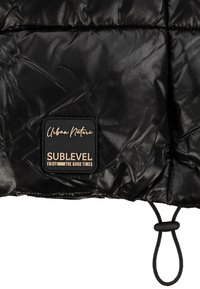 Schwarze Steppjacke mit glänzendem Finish, versehen mit einem quadratischen Etikett mit dem Schriftzug „Urban Nature“ und „SUBLEVEL“ sowie einem Kordelzug am Saum.