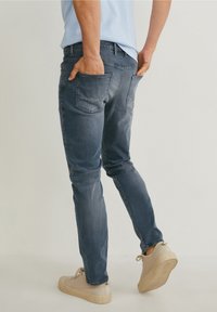 Slim-fit mörkblå jeans med bakfickor, visade från sidan. Bärs med beiga sneakers. Lätt tygtextur, avslappnad design.