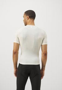 T-shirt de sport blanc, ajusté, à manches courtes, avec des coutures minimales et un col arrondi. Porté avec un pantalon de sport noir, mettant en valeur une texture lisse.
