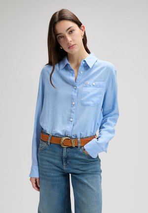 Jeune femme aux longs cheveux bruns portant une chemise bleu clair à boutons, un jean bleu et une ceinture marron, posant avec une main dans la poche.