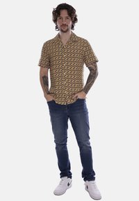 Camicetta a maniche corte marrone con un motivo circolare, abbinata a jeans blu e sneakers bianche. Tatuaggi notevoli visibili sulle braccia.