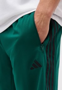 Χέρι στην τσέπη πράσινου φόρμα Adidas με μαύρες ρίγες και τσέπη με φερμουάρ, φορώντας λευκό πουκάμισο.