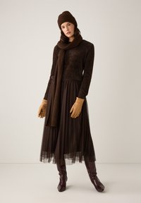 Braune Strickmütze, Schal und Handschuhe kombiniert mit einem flauschigen dunkelbraunen Oberteil und einem plissierten Tüllrock. Das Model trägt glänzende burgunderfarbene Stiefel.