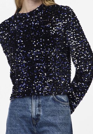 Czarna bluzka z długim rękawem pokryta niebieskimi i srebrnymi cekinami, noszona z niebieskimi dżinsami, widoczna od barków do talii.