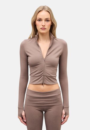 Mujer de pie, mirando al frente, vistiendo una camiseta corta de manga larga con cremallera en color topo y leggings a juego de cintura alta, sobre un fondo liso.