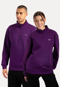 Sudaderas de cuello con cremallera en morado, fabricadas en un tejido suave, con puños y dobladillo acanalados, y un pequeño logo de la marca en el pecho.