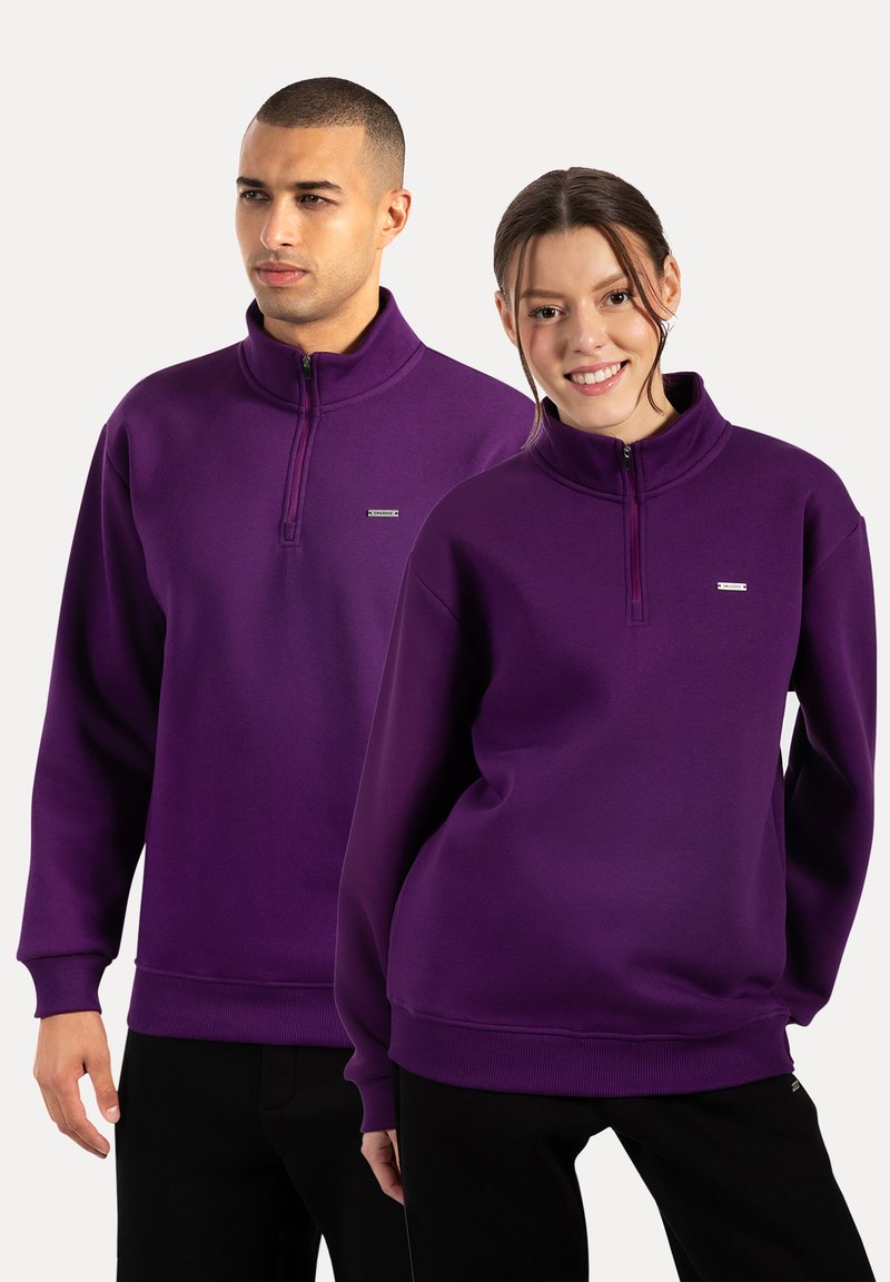 Sudaderas de cuello con cremallera en morado, fabricadas en un tejido suave, con puños y dobladillo acanalados, y un pequeño logo de la marca en el pecho.