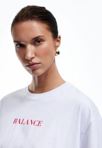 T-shirt blanc avec le mot "BALANCE" en rouge, associé à de grandes boucles d'oreilles en or, mettant en valeur une texture de tissu lisse et une coupe décontractée.