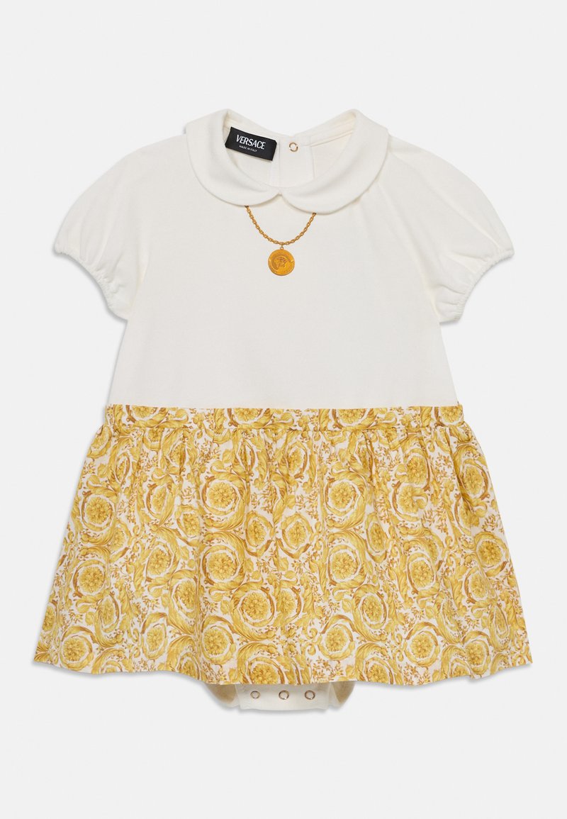 Vestido branco de manga curta com uma saia de padrão floral amarelo, apresentando um colarinho e um detalhe de medalhão dourado na gola.
