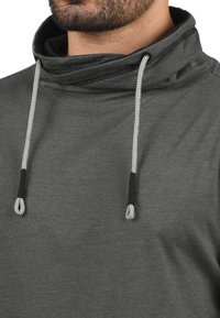 Grijze hoodie met een hoge kraag, voorzien van een gestructureerde stof, zwarte trekkoorden en zilveren agletdetails.