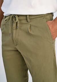 Olijfgroene broek met een trekkoord in de taille, slim fit en zijzakken. De stof lijkt een gestructureerde textuur te hebben met een gladde afwerking en subtiele plooien.