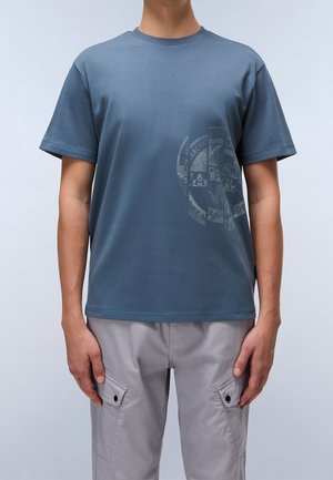 Mand iført en blå T-shirt med grafik og lysegrå cargobukser, stående mod en ensfarvet lys baggrund.