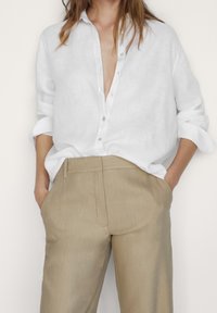 Camicia bianca in lino con maniche arrotolate, abbinata a pantaloni beige chiaro su misura con una texture liscia e linee pulite.